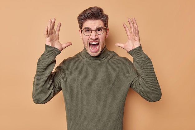 Free photo photo of outraged man yells from annoyance screams loudly keeps palms raised expresses negative emotions wears round spectacles and sweater isolated over beige background furious male model
