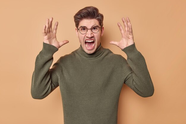 Photo of outraged man yells from annoyance screams loudly keeps palms raised expresses negative emotions wears round spectacles and sweater isolated over beige background Furious male model