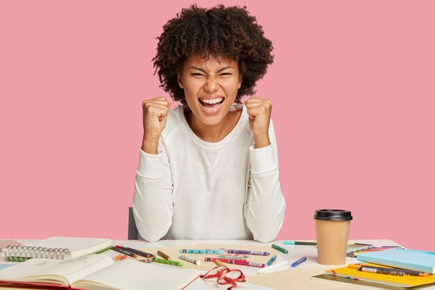 Photo of overjoyed African American architect keeps fists clenched, smiles broadly, squints face