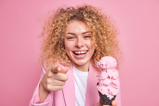 大喜びの縮れ毛の女性が喜んで笑う写真は、食欲をそそるアイスクリームの笑顔を直接保持し、ピンクの壁に白い歯が孤立していることを広く示しています。不健康な食事の概念