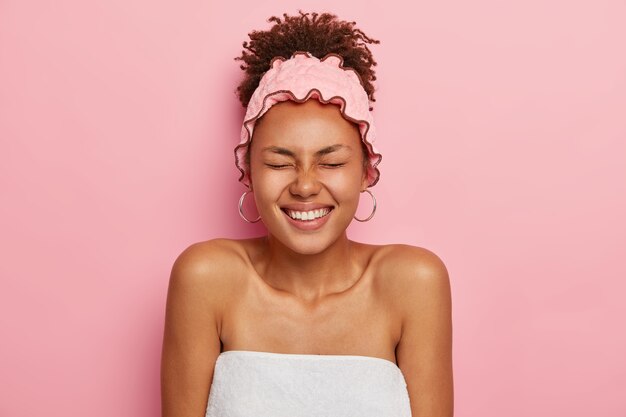 Photo of overjoyed dark skinned woman wrapped in white bath towel, wears pink shower headband, preapres for sauna, has healthy well cared dark skin, curly combed hair, expresses good feelings