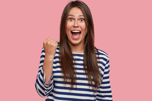 Photo of overjoyed woman feels like champion, shows victory gesture, raises clenched fists