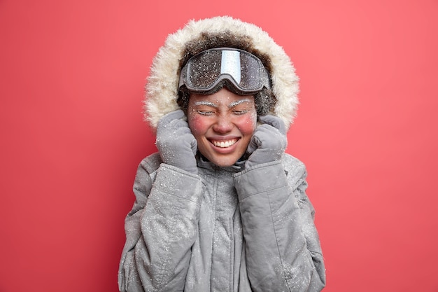 Photo of overjoyed woman wears hood of grey jacket smiles pleasantly has red face covered with frost goes skiing during December.