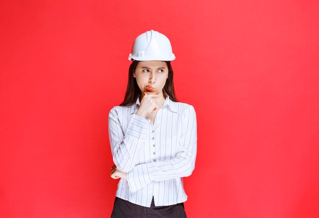 赤い壁に立っている安全帽子をかぶった物思いにふけるビジネス女性の写真。