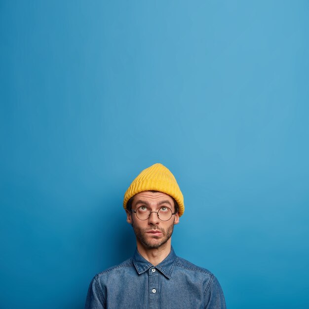 Фотография задумчивого сосредоточенного мужчины задумчиво смотрит вверх, в желтой шляпе, джинсовой рубашке, круглых больших очках, стоит над синей стеной, копирует пространство вверх.