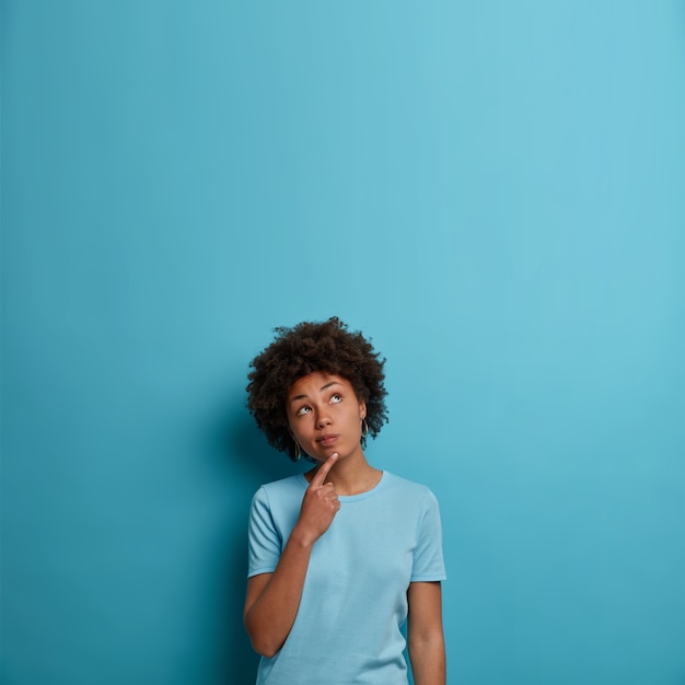 Photo of pensive dark skinned woman keeps finger on chin, concentrated above and ponders about something, wears t shirt, poses against blue wall, empty space for your promotion or information