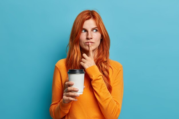 Photo of pensive good looking ginger woman keeps finger on lips and thinks deeply about something makes plans for tomorrow holds take away coffee. Beautiful red haired female drinks tea indoor