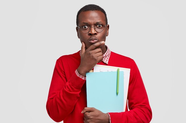 Photo of perplexed indignant black man holds chin, carries folders and papers, stares  with stupefied expressions