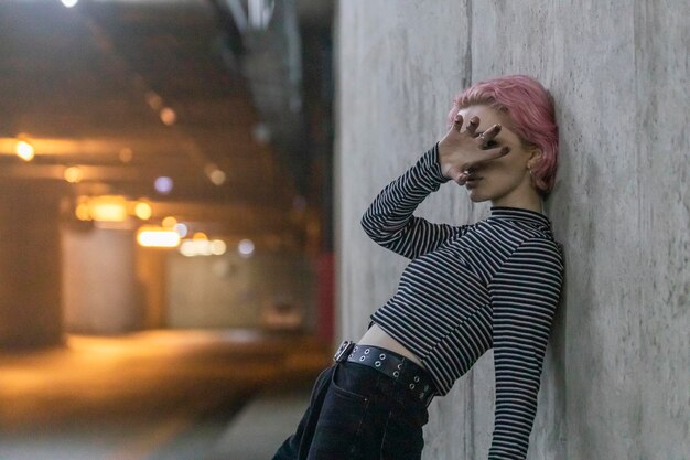 通りの壁に寄りかかって彼女の顔に手を置いたピンクの髪の少女の写真