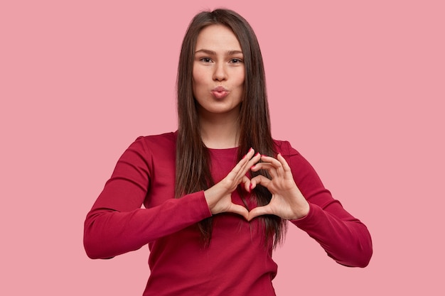 Photo of pleasant looking woman makes lips round, shows heart gesture with fingers, flirts with boyfriend on distance, has appealing appearance