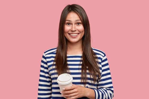 Photo of pleased brunette girl dressed in casual striped sweater, drinks takeaway coffee