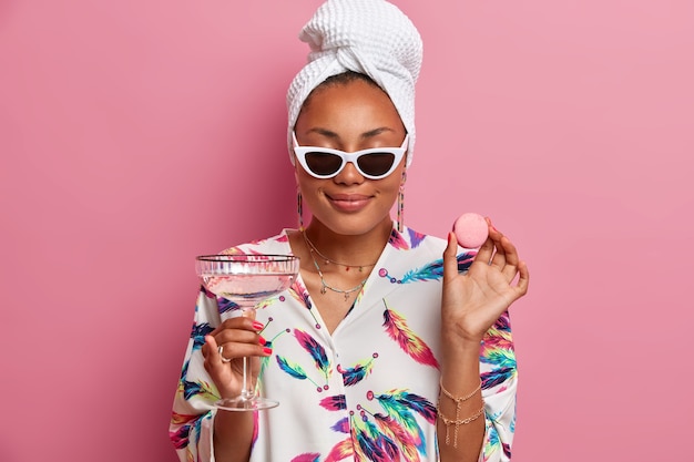 Photo of pleased dark skinned woman closes eyes smiles gently enjoys spending time at home holds delicious macaroon and glasses of martini cocktail wears dressing gown isolated over pink wall