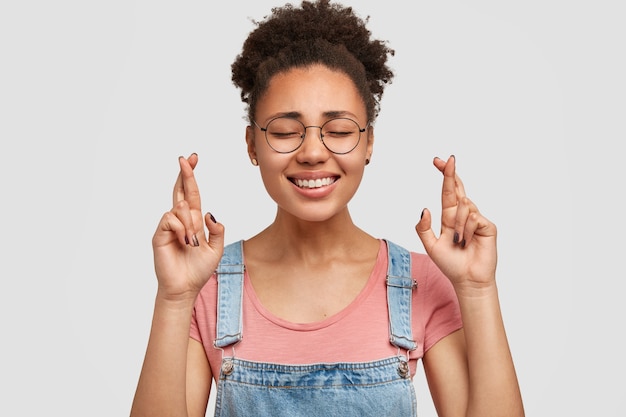 Photo of pleased dark skinned young female with happy expression, crosses fingers, believes in good luck, has toothy smile, poses against white wall. Body language concept. Positiveness