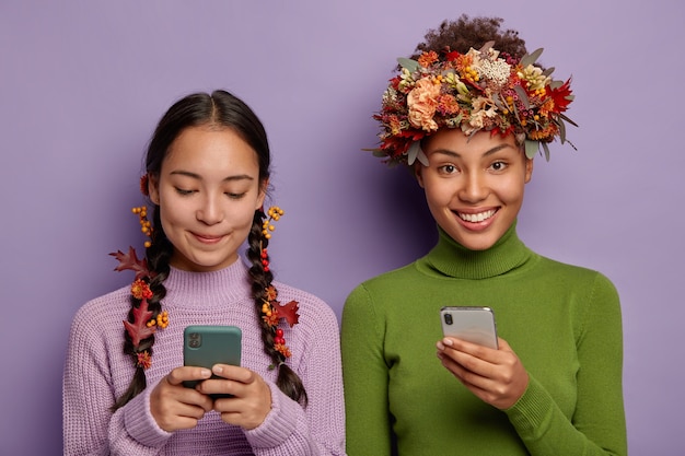 Photo of pleased female friends hold modern smartphones, send messages to friends, use creative way to wear autumn leaves, have glad expressions, wears sweaters.