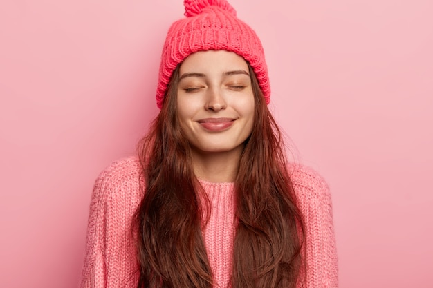 Photo of pleased long haired pleased Caucasian woman keeps eyes closed, smiles gently, has healthy skin, wears warm knitted hat and jumper, poses against pink background, imagines something nice