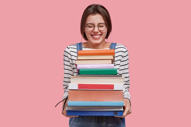 Photo of pleased teenage girl holds heap of textbooks, being in high spirit, wears denim overalls, poses against pink background