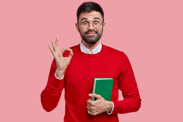 Photo of pleased unshaven man makes okay gesture, looks joyfully, has dark stubble, says everything is alright, holds notebook