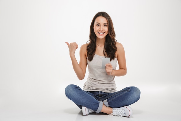 Photo of pleased woman with long brown hair sitting with legs crossed on the floor using silver smartphone and pointing away, isolated over white wall