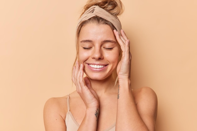 Free photo photo of pleased young european woman keeps eyes closed touches face gently has natural beauty healthy clean skin wears headband has combed hair isolated over beige background. wellness concept