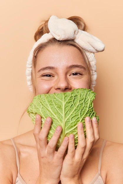 Photo of pleased young woman covers mouth with leaf of cabbage uses fresh vegetable for making natural cosmetic product has well cared healthy skin wears headband isolated on beige background