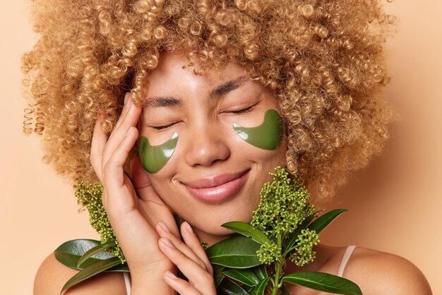 Photo of pleased young womann with curly bushy hair keeps hand on face smiles happily has eyes closed holds green plant applies beauty patches under eyes uses natural cosmetic Skin care concept