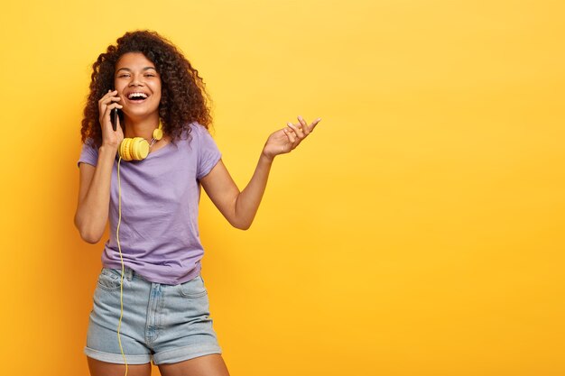 Photo of positive African American girl talks on mobile phone, smiles broadly, raises hand, shares impressions about shopping, discusses last fashion trends