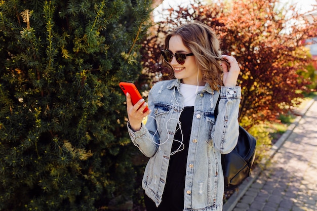 Photo of a positive cheerful teenage girl spends time in the park and using mobile phone.