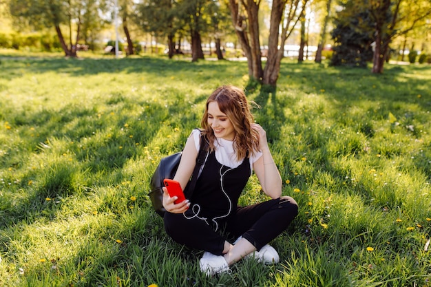 Photo of a positive cheerful teenage girl spends time in the park and using mobile phone.