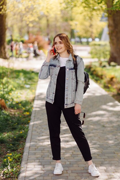 ポジティブで陽気な10代の少女の写真は、公園で携帯電話を使用して時間を過ごします。