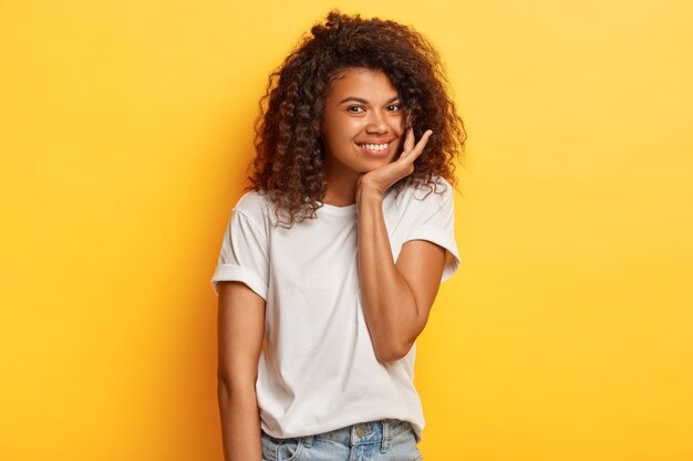 Photo of positive dark skinned woman with crisp hair, has gentle smile, touches chin, dressed in casual white t shirt and jeans, stands against yellow wall.
