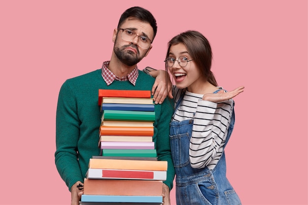 Photo of positive European woman wears striped sweater and overalls, leans at shoulder of tired male nerd with pile of thick books