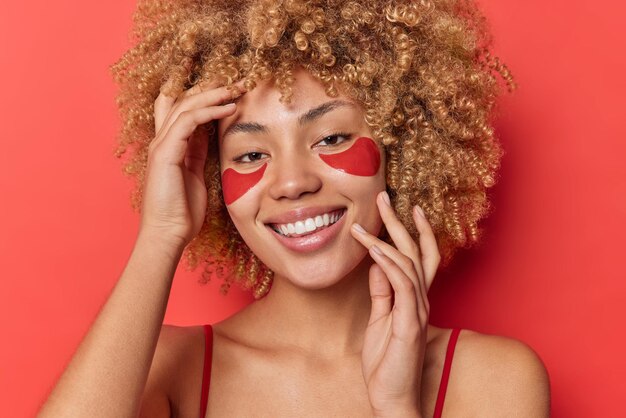 Free photo photo of positive woman with curly blonde hair applies collagen patches under eyes for reducing wrinkles and fine lines smiles toothily isolated over red background. skin care and wellness concept