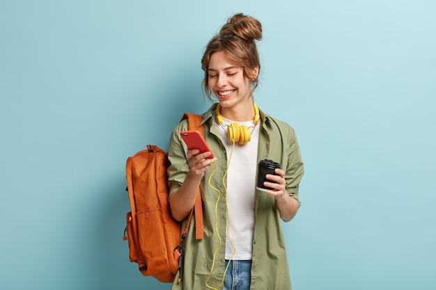 캐주얼 옷에 긍정적 인 젊은 여성의 사진, 휴대 전화에 멀티미디어 파일 다운로드, 목에 헤드폰 있음