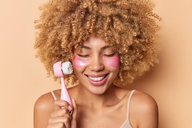 Photo of positive young woman with curly bushy hair uses face massager applies hydrogel patches under eyes for skin treatment smiles happily keeps eyes closed isolated over beige background.