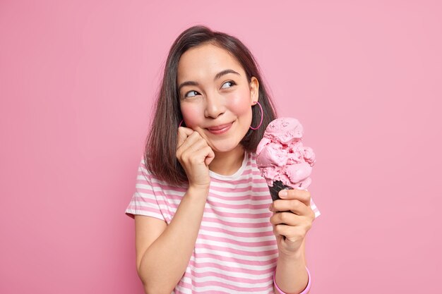 Photo of pretty Asian Woman looks dreamily aside recalls pleasant memories eats tasty summer dessert holds big cone ice cream dressed in casual t shirt models