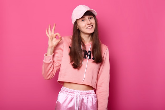 Photo of pretty attractive lady wearing rosy sport pullover, sweatpants, cap, shows okay sign