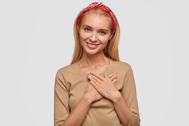 Photo of pretty blonde smiling young woman with appealing look, keeps hands on chest, expresses good feeling