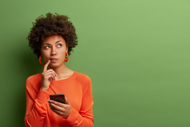 Photo of pretty ethnic woman ponders on how to answer question, thinks deeply about something, uses modern mobile phone, tries to made up good message, keeps index finger near lips, stands indoor