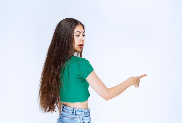 Photo of a pretty girl model standing and pointing aside against white wall.