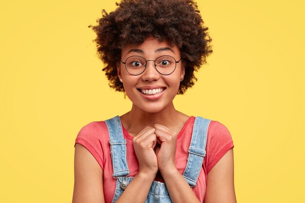 Photo of pretty happy dark skinned female with pleasant smile, keeps hands together, recieves good words of praise, has curly hair, poses against yellow wall. Glad African American woman