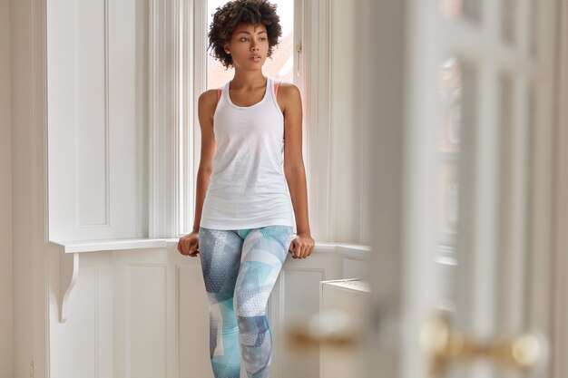 Photo of pretty relaxed fitness trianer rest at home after training, wears casual white vest and colorful leggings