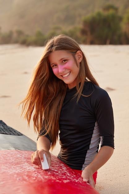 Photo of pretty surfer in high spirit, dressed in wetsuit, has toothy smile, long hair, waxes surfaces of board for hiting ocean waves