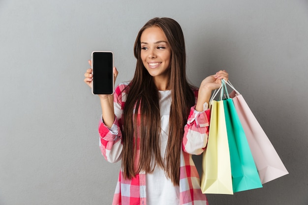 Photo of pretty woman holding shopping bags, showing blank mobile screen