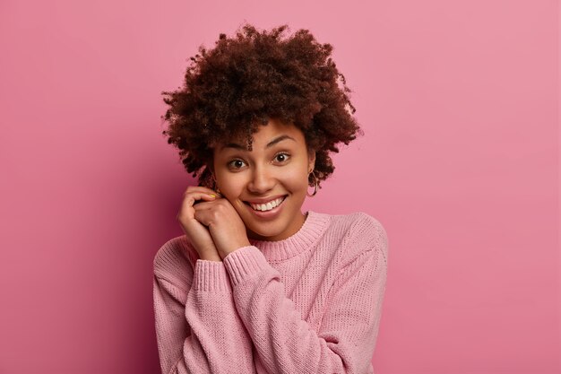 Photo of pretty young tender woman with Afro hair smiles gently, keeps hands near face, looks directly , wears casual sweater, hears somethning good, poses against pink  wall