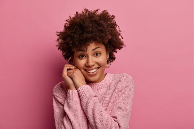 На фото симпатичная молодая нежная женщина с афро-волосами нежно улыбается, держит руки возле лица, смотрит прямо, носит повседневный свитер, слышит что-то хорошее, позирует у розовой стены