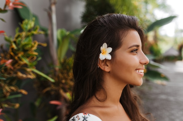 Photo in profile of young positive woman with tanned skin and flower in dark hair posing against wall of tropical plants