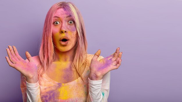 Photo of puzzled European female showered with colorful powder, spends weekend on Holi festival, spreads hands, keeps mouth opened, models over violet wall, blank space for your text.