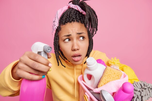 Photo of puzzled mixed race woman holds detergent for washing windows basket with cleaning supplies