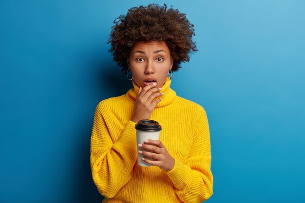 Photo of puzzled scared dark skinned young woman holds chin, poses with takeaway coffee isolated over blue wall