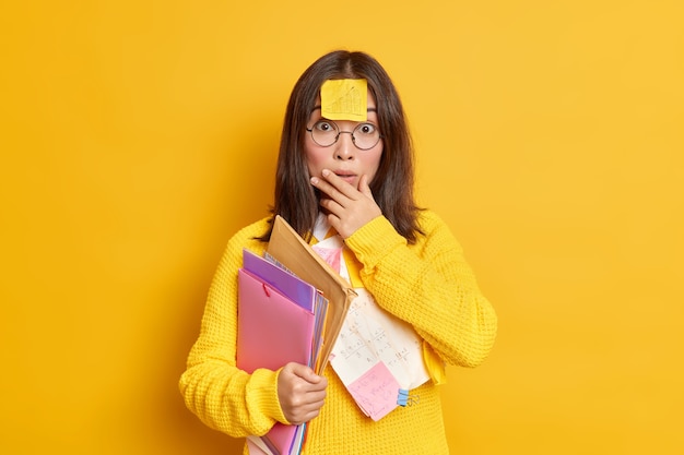 Free photo photo of puzzled surprised asian female student has memo note stuck on forehead prepares coursework carries folders with papers makes education project works at her assignment studies remotely